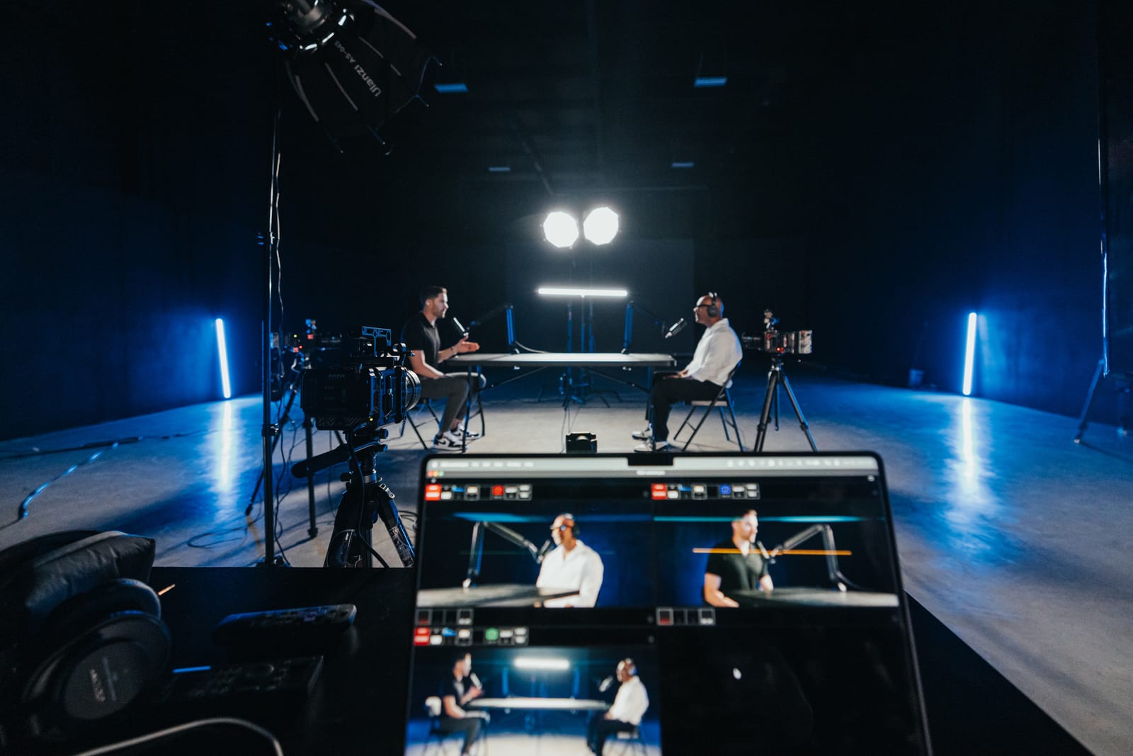 Two hosts recording a podcast at a table, three cameras running, the multicam feed on a monitor in the foreground